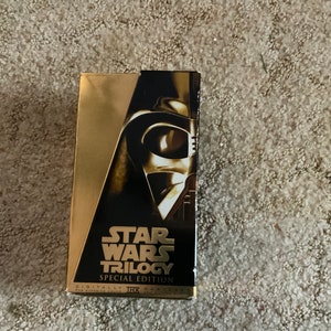Pode incluir: Uma fita VHS Star Wars Trilogy Special Edition dourada e preta com o capacete de Darth Vader. A caixa VHS tem detalhes dourados e o logotipo de Star Wars. O texto "Digitally Mastered for Superior Sound" é visível.