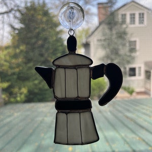May include: A white and black stained glass coffee pot hanging from a window with a suction cup.