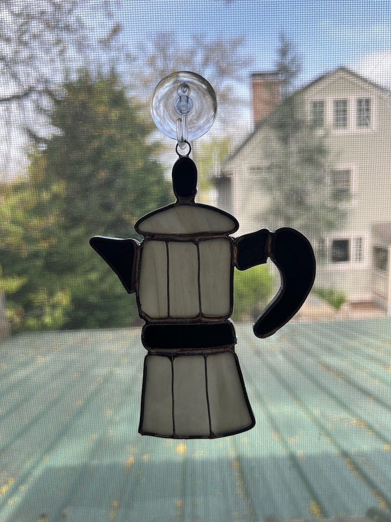 May include: A white and black stained glass coffee pot hanging from a window with a suction cup.