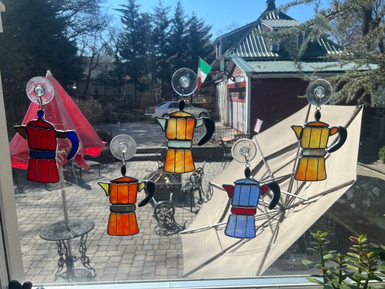 May include: Five stained glass coffee pot ornaments hanging in a window. The ornaments are red, orange, yellow, blue, and white. Each ornament has a suction cup for hanging.
