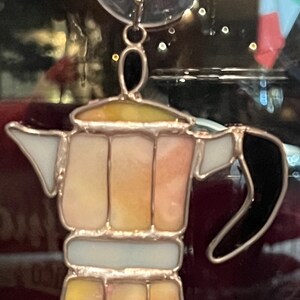 May include: A stained glass coffee pot with a black handle and a yellow, orange, and white design. The coffee pot is hanging from a suction cup.