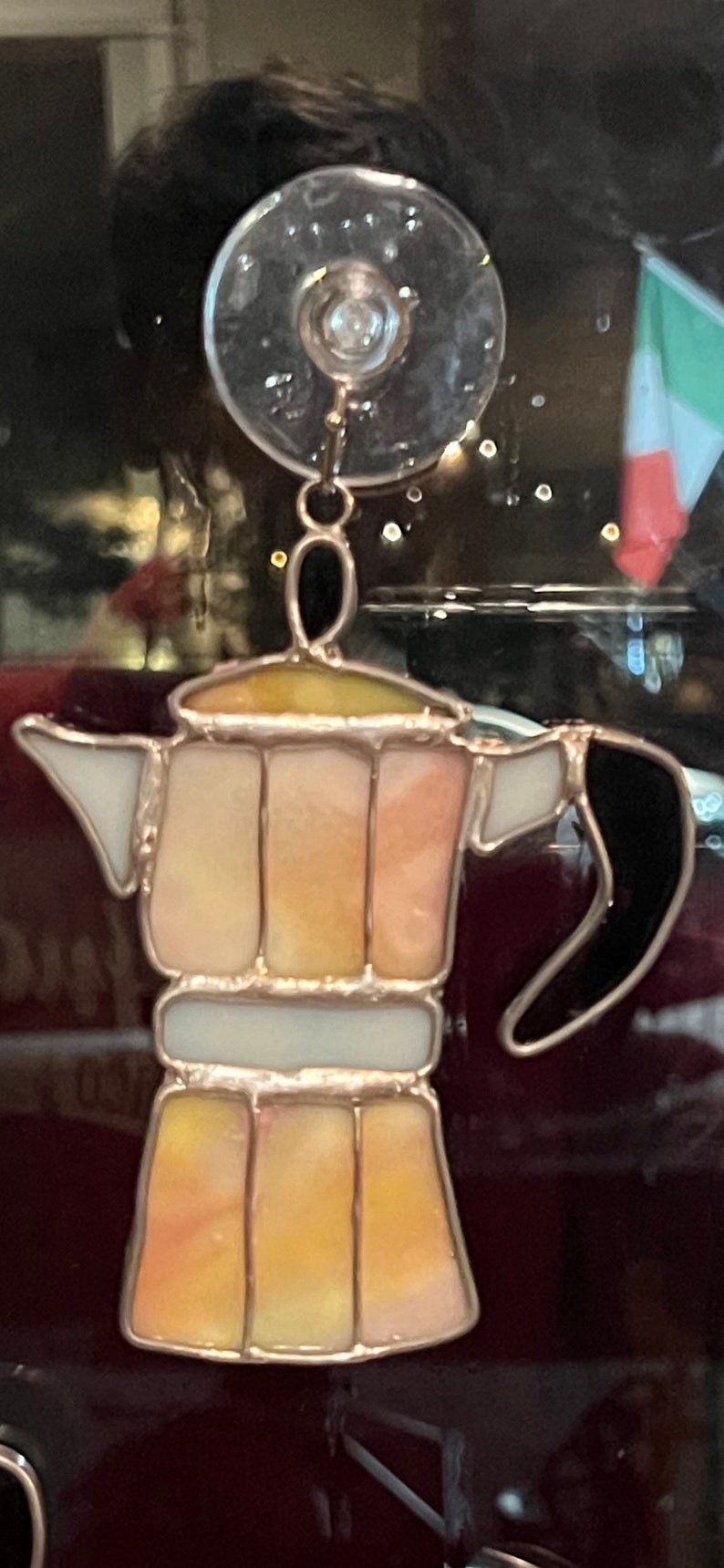 May include: A stained glass coffee pot with a black handle and a yellow, orange, and white design. The coffee pot is hanging from a suction cup.