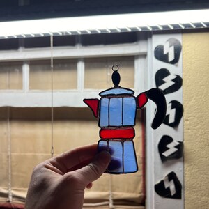 May include: A stained glass window hanging ornament shaped like a blue and red coffee pot.