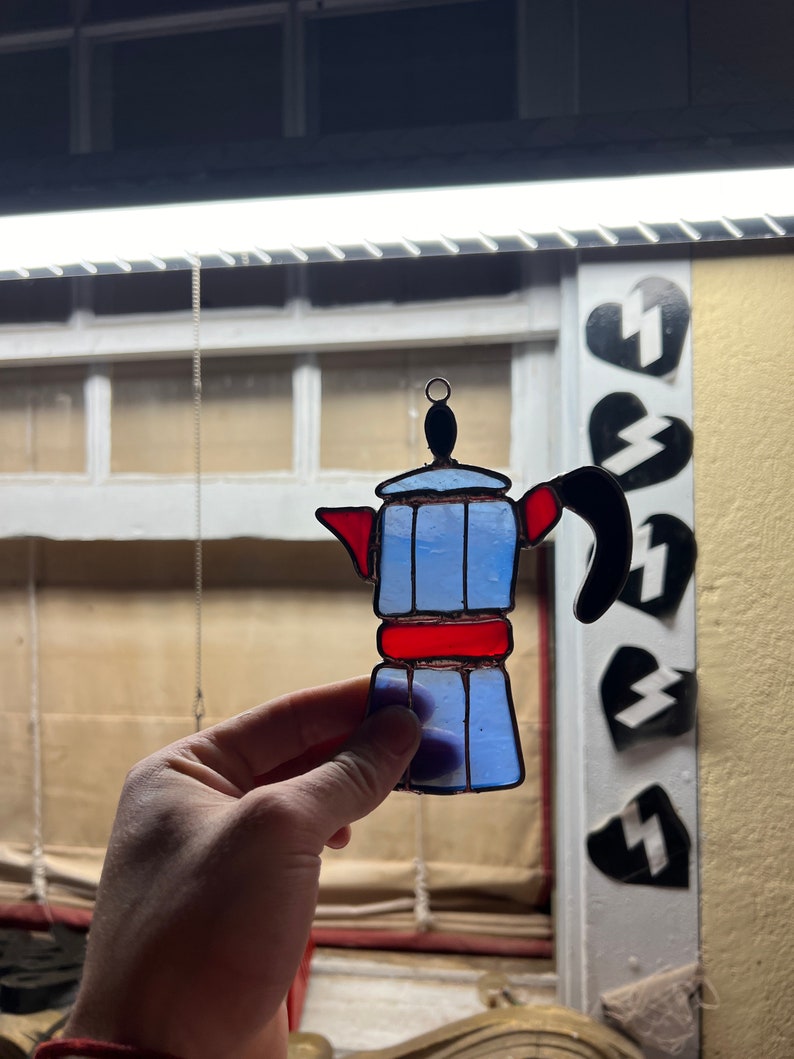 May include: A stained glass window hanging ornament shaped like a blue and red coffee pot.
