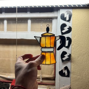 May include: A stained glass coffee pot ornament in shades of yellow and orange. The ornament is hanging from a chain and is being held by a hand.