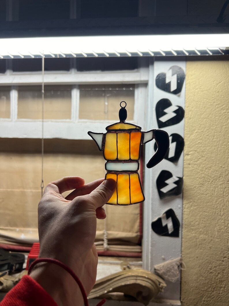 May include: A stained glass coffee pot ornament in shades of yellow and orange. The ornament is hanging from a chain and is being held by a hand.