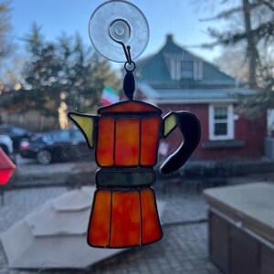 May include: A stained glass coffee pot with a suction cup for hanging on a window. The coffee pot is orange, yellow, and black. The background is blurry and shows a house and trees.