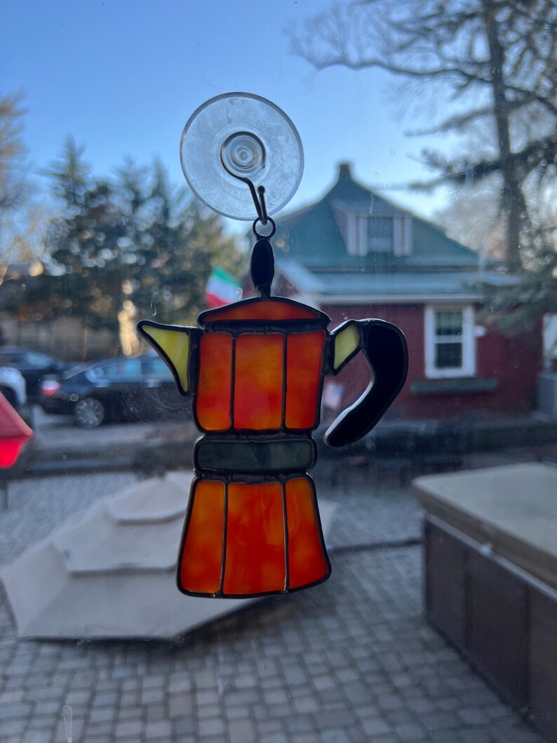 May include: A stained glass coffee pot with a suction cup for hanging on a window. The coffee pot is orange, yellow, and black. The background is blurry and shows a house and trees.