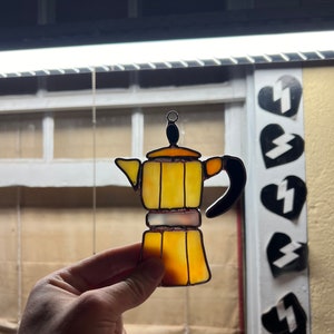 May include: A yellow stained glass Moka pot with a black handle. The pot is hanging from a chain and is in front of a window.