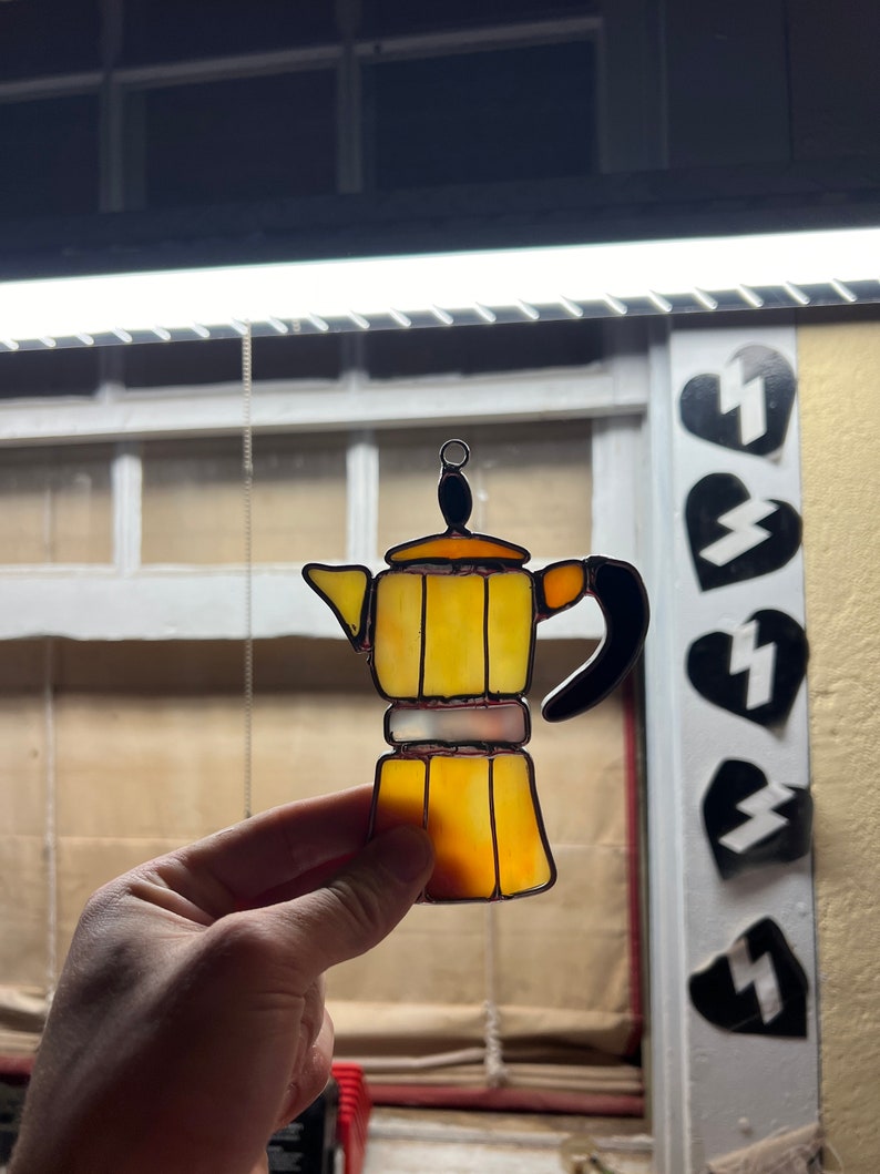 May include: A yellow stained glass Moka pot with a black handle. The pot is hanging from a chain and is in front of a window.