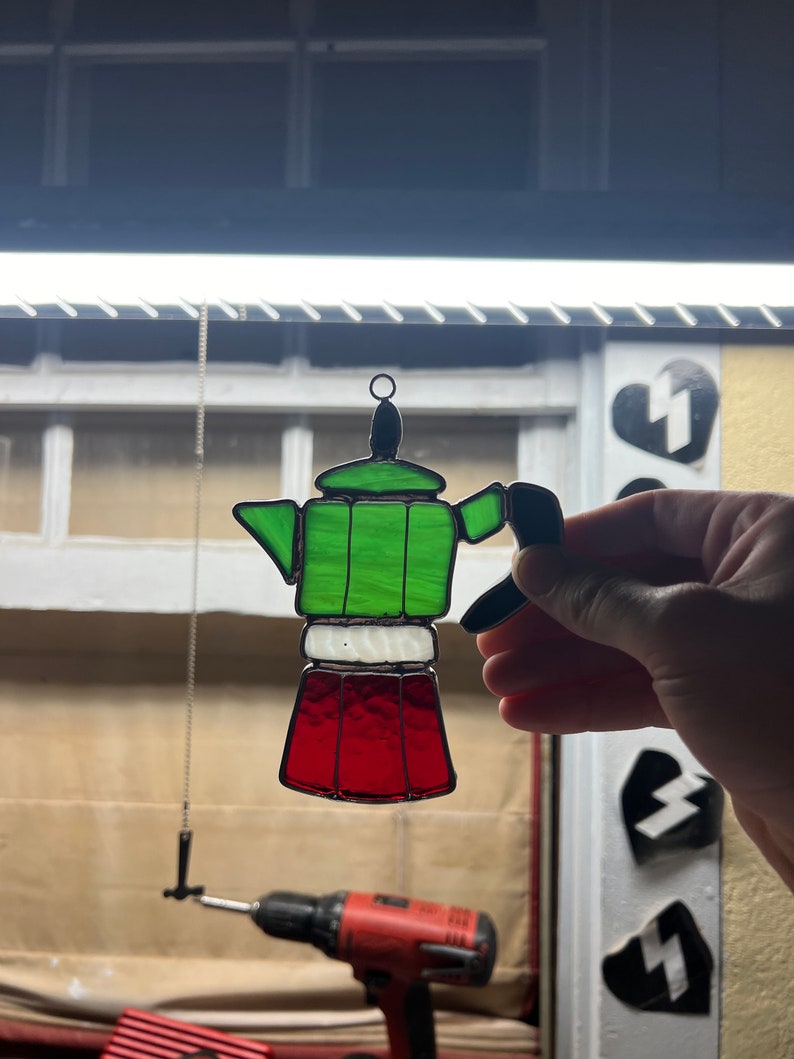 May include: A stained glass hanging ornament shaped like a green, white, and red Italian coffee pot.