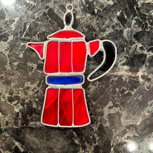 May include: A red and black stained glass coffee pot ornament. The coffee pot has a silver trim and a blue band around the middle.