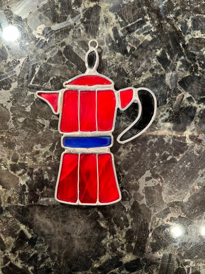 May include: A red and black stained glass coffee pot ornament. The coffee pot has a silver trim and a blue band around the middle.