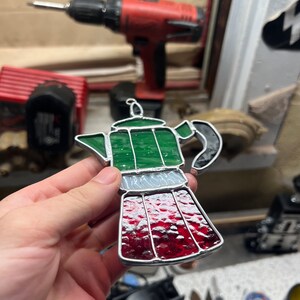 May include: A stained glass depiction of a green and red coffee pot. The coffee pot has a silver band around the middle and a black handle.