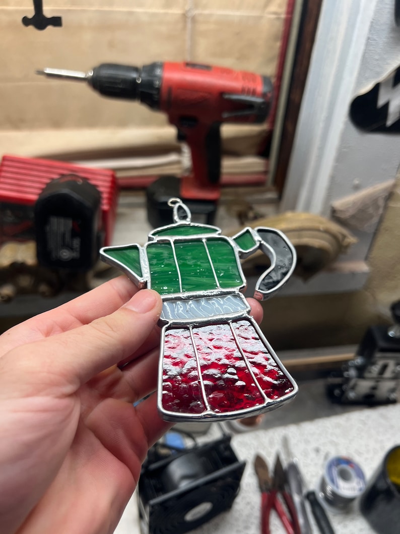 May include: A stained glass depiction of a green and red coffee pot. The coffee pot has a silver band around the middle and a black handle.