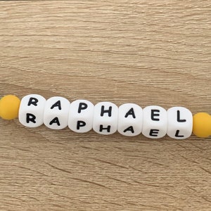 May include: A blue silicone baby teether with a bear on each end. The teether has a clip on one end and a string on the other. The beads are yellow, turquoise, and white with the name "RAPHAEL" spelled out on the white beads.