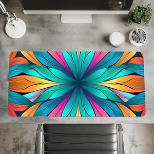 Op de afbeelding: Een rechthoekige bureaumat met een levendig bloemenontwerp. Het patroon heeft teal, oranje, roze en gele bloembladachtige vormen die uitstralen vanuit een centraal punt. De mat ligt op een grijze bureau met een computermonitor en accessoires.