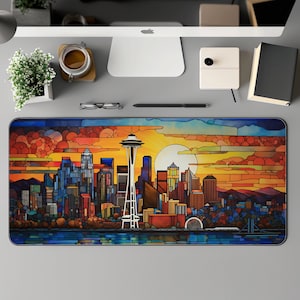 May include: A rectangular desk mat featuring a colorful stained-glass style depiction of the Seattle skyline at sunset. The Space Needle is prominently displayed, with the sun setting behind the city. The mat is on a gray desk with other office supplies.