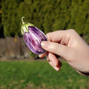 May include: A watercolor painting of an eggplant. The eggplant is a deep purple color with white stripes and a green stem. The painting is held in a hand against a blurred background of green foliage.
