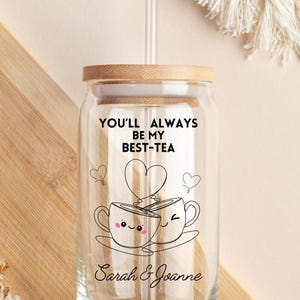 May include: A clear glass reusable cup with a bamboo lid and a straw. The cup features a black and white illustration of two teacups with hearts and the text "You'll always be my best-tea". The cup is personalised with the names "Sarah & Joanne".