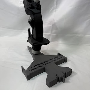 Op de afbeelding: Een zwarte joystick met een grijze basis in de vorm van een F-16 straaljager. De joystick heeft verschillende knoppen en een rode knop. De basis heeft de tekst "F-16 FIGHTING FALCON 138TH FW, 125TH FS "Tulsa Vipers".