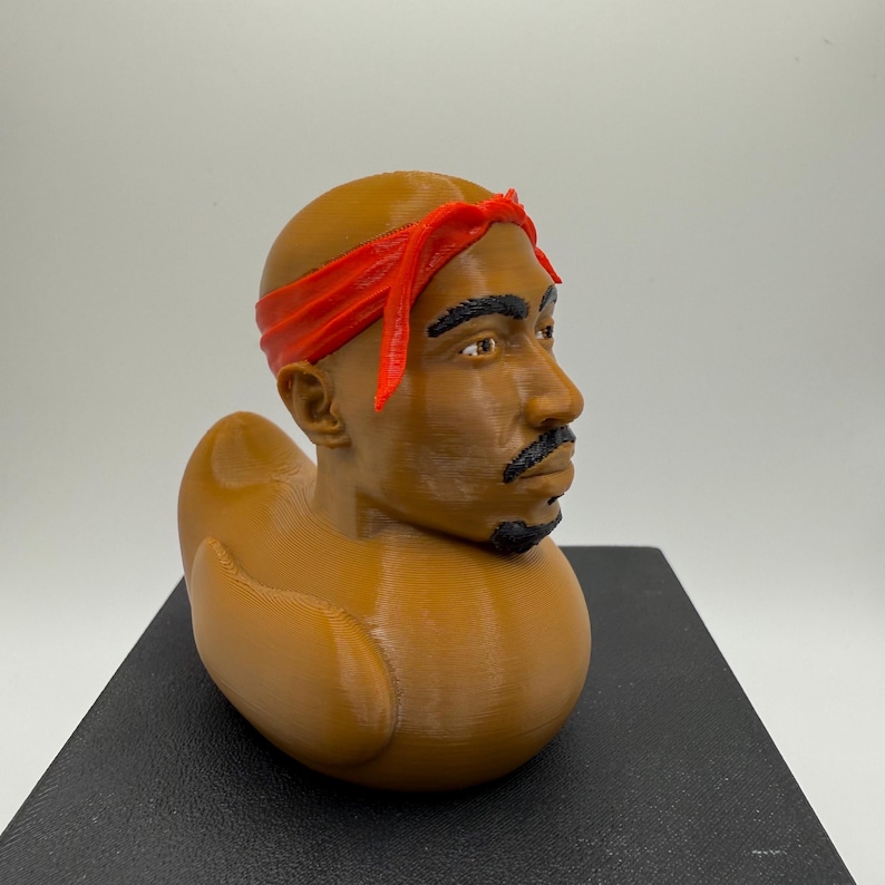 May include: A brown novelty duck figurine with a red bandana. The duck's head is a detailed bust of a man with a mustache and goatee. The figurine is resting on a black surface.