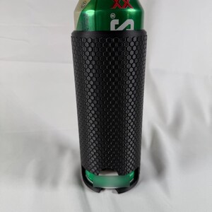 May include: A black, textured can cooler with a honeycomb pattern, holding a green and gold can. The cooler has a cylindrical shape and a green base, designed to keep beverages cold.