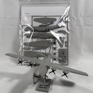 Puede incluir: Maqueta gris de un avión AC-130 Spooky con cuatro hélices por ala. El modelo está sobre un soporte con el texto "AC-130 Spooky". Las piezas de montaje son visibles en una bolsa de plástico transparente.