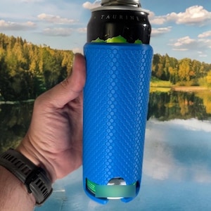 May include: A blue can cooler with a honeycomb pattern, holding a black and silver can. The cooler is cylindrical and has a textured surface. The can has the word "TAURINE" printed on it. The background shows a lake and trees.