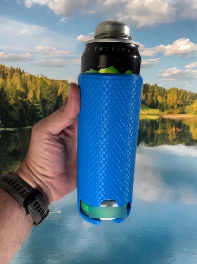 May include: A blue can cooler with a honeycomb pattern, holding a black and silver can. The cooler is cylindrical and has a textured surface. The can has the word "TAURINE" printed on it. The background shows a lake and trees.