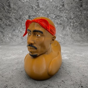 May include: A brown novelty duck figurine with a sculpted head of a man wearing a red bandana. The duck has a detailed face with a mustache and beard. The figurine is on a gray surface.