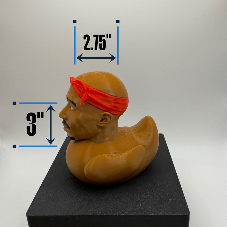 May include: A brown rubber duck with a 3-inch height and 2.75-inch width, featuring a sculpted head with a red bandana. The duck is resting on a black surface, showcasing a unique and artistic design.