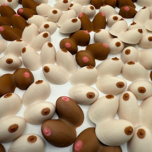 Puede incluir: Una colección de pequeños senos esculpidos en varios tonos de piel, incluyendo marrón claro y oscuro, y blanco roto. Cada seno tiene un pezón con una areola rosa. Los objetos están dispuestos juntos sobre una superficie blanca.