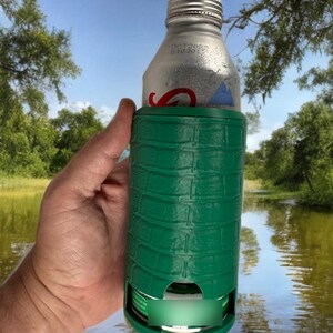 May include: A green insulated beverage holder with a textured, crocodile-like pattern. The holder is wrapped around a silver aluminum bottle. The background shows a lake and trees.