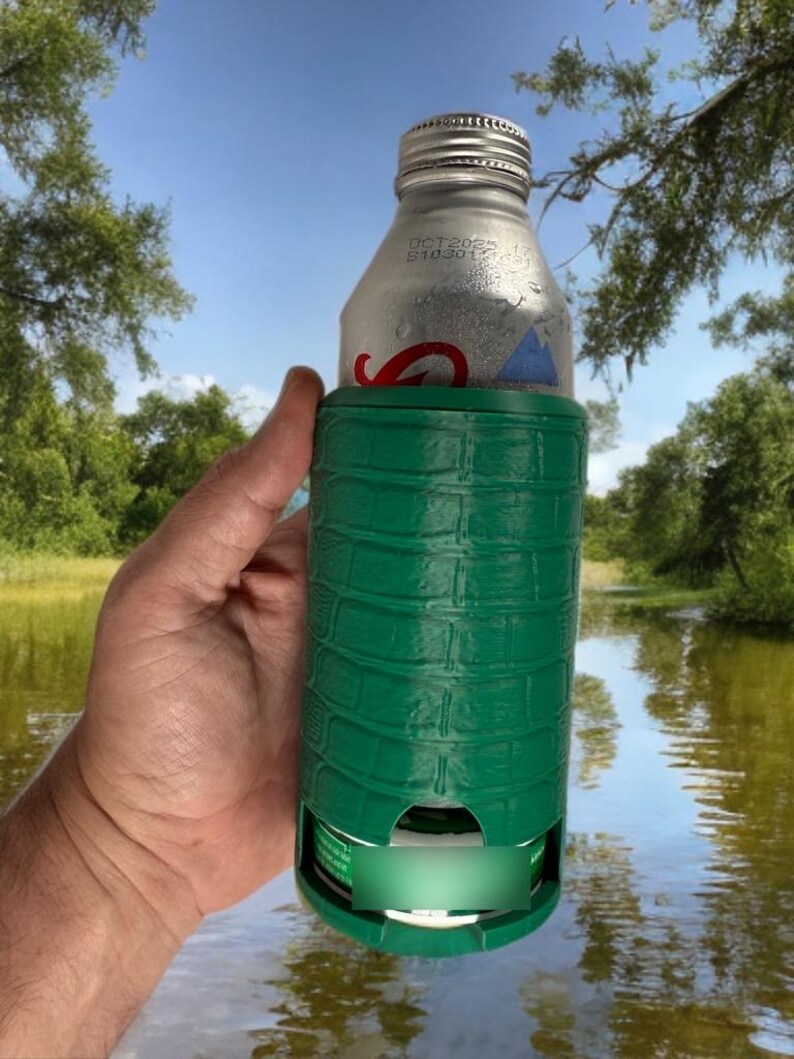May include: A green insulated beverage holder with a textured, crocodile-like pattern. The holder is wrapped around a silver aluminum bottle. The background shows a lake and trees.