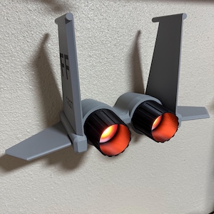 Puede incluir: Un modelo de avión gris con el texto "FF" y "1027" en la cola. Los motores a reacción tienen una luz naranja brillante. El modelo está montado en una pared blanca.