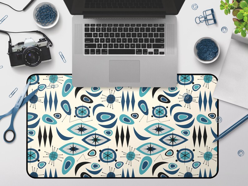 Op de afbeelding: Een bureauonderlegger met een retro modern design uit het midden van de eeuw, met een herhalend patroon van blauwe, turquoise en zwarte abstracte vormen op een cr&egrave;me achtergrond.