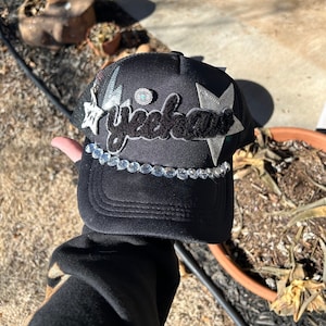 Puede incluir: Gorra de camionero negra con un parche negro "yeehaw", parches de estrellas plateadas y una piedra turquesa. La gorra está decorada con una fila de diamantes de imitación transparentes.