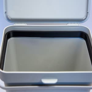 Könnte beinhalten: Ein weißer Mülleimer mit einer schwarzen Gummidichtung am oberen Rand. Der Deckel ist offen und der Innenraum des Eimers ist leer.