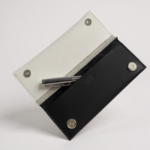 Puede incluir: Un estuche para bolígrafo de cuero blanco y negro con un bolígrafo plateado en su interior. El estuche está abierto, revelando un interior blanco y un bolsillo negro. El estuche se asegura con cierres de presión plateados. La palabra "KAMI" está grabada en el bolsillo negro.