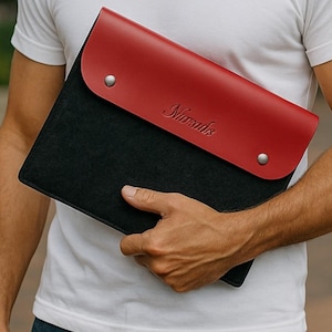 May include: A black and red leather laptop sleeve with the word "Nomads" embossed on the red flap. The sleeve is held by a person wearing a white t-shirt. The sleeve has two silver button closures.