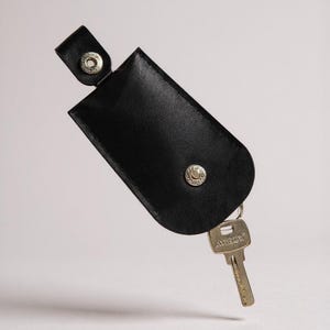 Könnte beinhalten: Schwarzer Leder-Schlüsselhalter mit silbernem Druckknopfverschluss und einem einzelnen Schlüssel. Der Schlüssel ist silberfarben und trägt das Wort "Artec". Der Schlüsselhalter ist rechteckig mit abgerundeten Kanten.