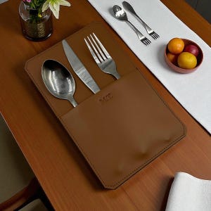 May include: A brown leather cutlery holder with a spoon, knife, and fork. The holder has the initials "M.T." embossed on the front. The cutlery is stainless steel. The holder is on a wooden table, with a white runner and a small bowl of fruit.