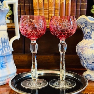 May include: Two ruby red crystal wine glasses with detailed cut patterns on a silver tray. The glasses have long, clear stems and round bases. A decorative jug and books are in the background.