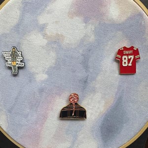 May include: An embroidery hoop with a fabric background featuring three enamel pins. One pin is a red football jersey with the number 87. Another pin has the text "Cause I was then I'd be the man". The third pin is a person with a braid.