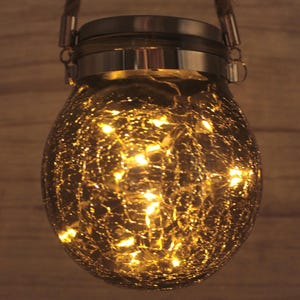 Puede incluir: Una linterna de vidrio dorada con un acabado craquelado y una tapa plateada. La linterna tiene un asa de cuerda y está iluminada con luces LED blancas cálidas.