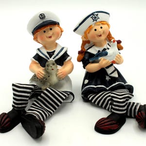 Könnte beinhalten: Zwei nautische Figuren von Kindern, eines hält eine Robbe und das andere einen Vogel. Sie tragen Matrosenmützen, marineblaue und weiße Outfits und gestreifte Socken. Die Figuren haben schwarze Schuhe mit roten Akzenten.