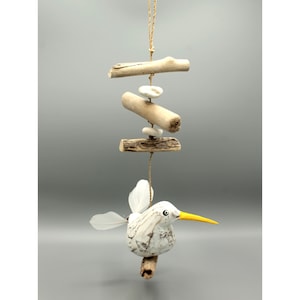 Può includere: Mobile in legno di deriva fatto a mano con un uccello intagliato, becco giallo e piume bianche. L'uccello è appeso sotto pezzi di legno di deriva e piccole pietre, sospeso da uno spago naturale. Decorazione ispirata alla natura.