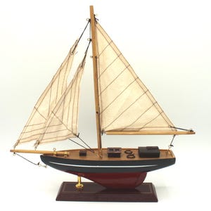 Può includere: Un modellino di barca a vela con scafo nero e una striscia bianca, chiglia rosso scuro e ponte in legno. Presenta due vele in tessuto color crema fissate ad alberi e boma in legno, con sartiame nero e piccole strutture marroni sul ponte.