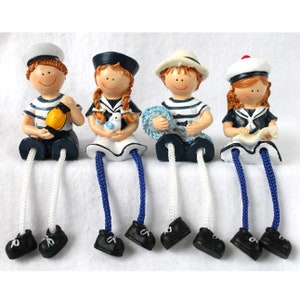 Può includere: Quattro figurine di marinai con camicie e cappelli a righe blu e bianche. Ogni figurina ha gambe lunghe e pendenti con una corda blu e bianca. Le figurine tengono vari oggetti nautici, tra cui un salvagente, un uccello, una corda e una mappa.